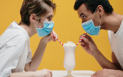 Two people wearing face masks trying to drink a milkshake through straws with a yellow background